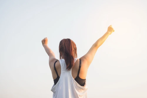 Woman in athletic wear raising arms against bright sky.