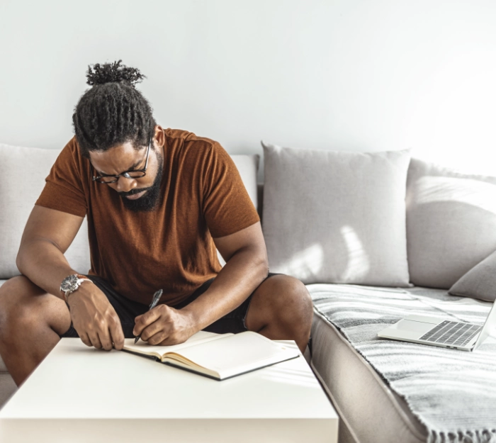 man writing in journal with laptop on sofa